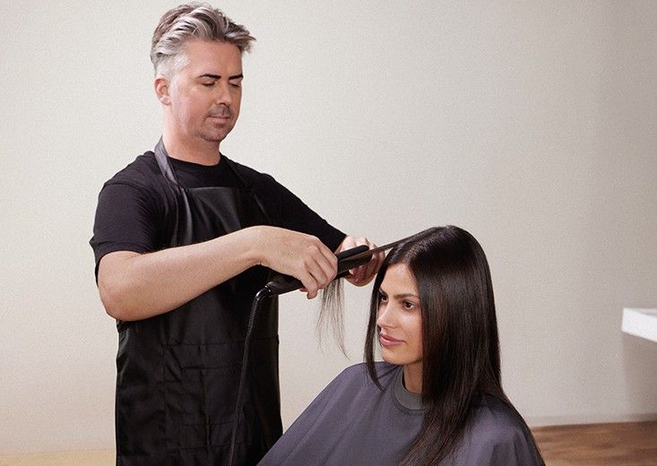 Kerasilk brand photo: a flat iron glides down a section of long, sleek dark hair on a seated client